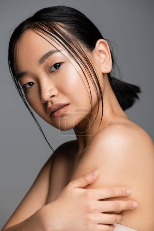 Photo for Portrait of brunette asian woman with natural makeup touching bare shoulder and looking at camera isolated on grey - Royalty Free Image