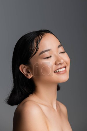 Photo for Young asian woman with bare shoulders and nude makeup smiling with closed eyes isolated on grey - Royalty Free Image