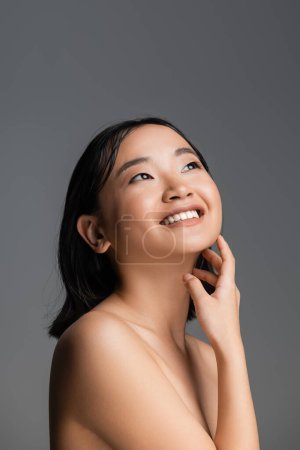 cheerful asian woman touching perfect skin and looking away isolated on grey