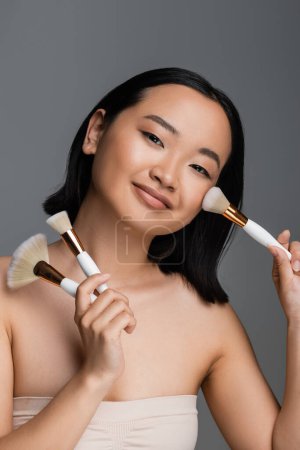 smiling asian woman holding cosmetic brushes and applying face powder isolated on grey