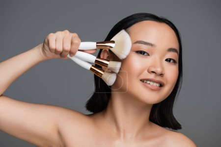 Photo for Happy asian woman with bare shoulders and natural makeup holding different cosmetic brushes isolated on grey - Royalty Free Image
