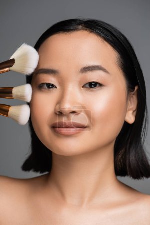 Téléchargez les photos : Portrait of brunette asian woman with natural makeup looking at camera near different cosmetic brushes isolated on grey - en image libre de droit