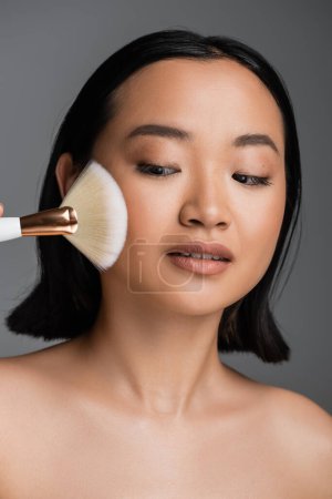 portrait of young asian woman with perfect skin applying face powder isolated on grey