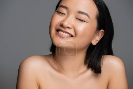 Photo for Portrait of smiling asian woman with naked shoulders and perfect skin smiling with closed eyes isolated on grey - Royalty Free Image