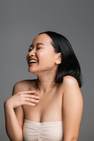 excited asian woman with closed eyes holding hand near chest and laughing isolated on grey