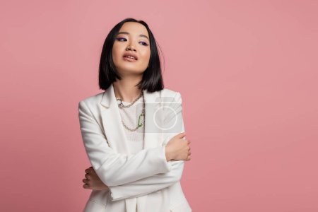 brunette asian woman in white jacket and necklaces standing with crossed arms and looking away isolated on pink