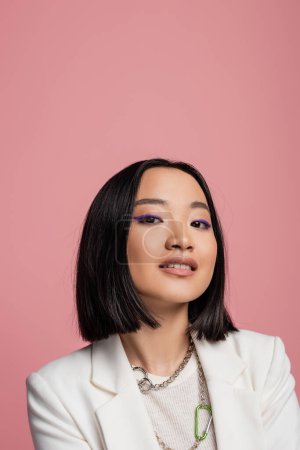 Photo for Portrait of brunette asian woman in makeup and white jacket looking at camera isolated on pink - Royalty Free Image