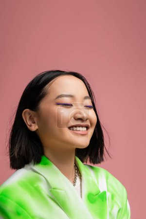 Photo for Portrait of pretty asian woman in green and white jacket smiling with closed eyes isolated on pink - Royalty Free Image