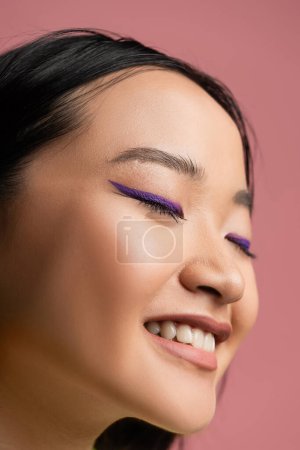 close up view of young asian woman with makeup and blue eyeliner smiling with closed eyes isolated on pink