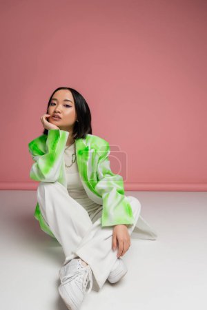 Photo for Full length of trendy asian woman in fashionable clothes and sneakers sitting on white surface and pink background - Royalty Free Image