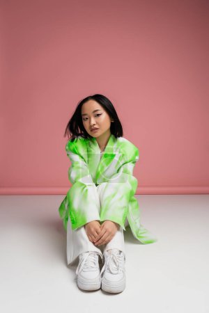 Foto de Full length of brunette asian woman in trendy clothes and sneakers sitting on white surface and pink background - Imagen libre de derechos