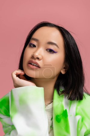 Photo for Portrait of asian woman in trendy clothes and makeup posing with hand near face isolated on pink - Royalty Free Image