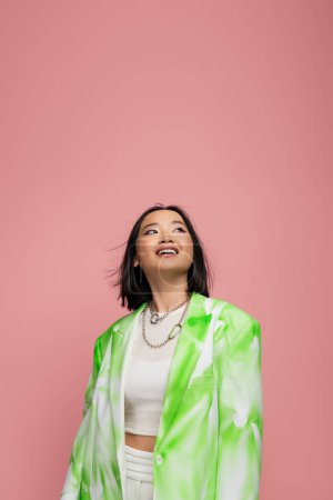 Photo for Happy asian woman in fashionable blazer and necklaces looking up isolated on pink - Royalty Free Image