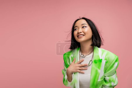 Téléchargez les photos : Stylish asian woman in blazer and metal necklaces touching chest and looking away isolated on pink - en image libre de droit