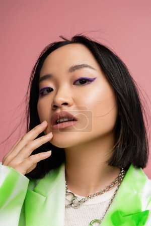 Photo for Portrait of stylish asian woman in silver necklaces touching face with makeup and looking at camera isolated on pink - Royalty Free Image