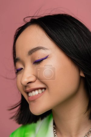 Photo for Close up portrait of brunette asian woman with blue eyeliner smiling with closed eyes isolated on pink - Royalty Free Image