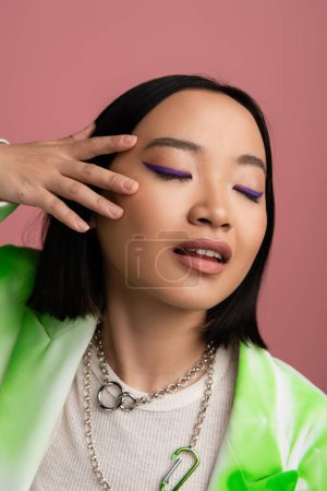 sensual asian woman in metal necklaces touching face with makeup and blue eyeliner isolated on pink