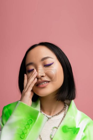 portrait of smiling asian woman with closed eyes touching perfect face with blue eyeliner isolated on pink