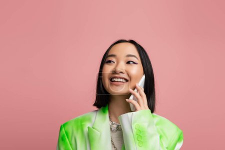 Photo for Fashionable asian woman smiling and looking away while talking on mobile phone isolated on pink - Royalty Free Image