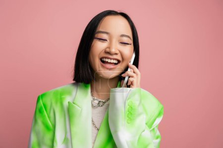 cheerful asian woman in white and green blazer talking on mobile phone isolated on pink