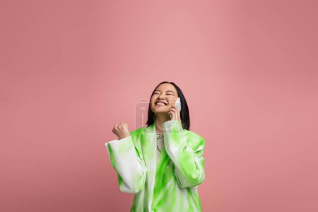 Foto de Excited asian woman in stylish white and green jacket talking on cellphone and showing win gesture isolated on pink - Imagen libre de derechos