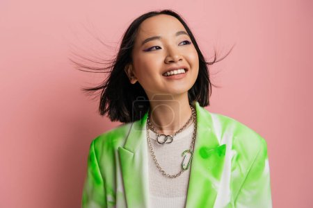 Photo for Joyful asian woman in trendy outfit and makeup with blue eyeliner looking away on pink background - Royalty Free Image