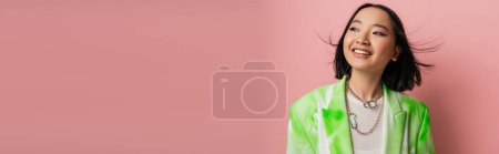 brunette asian woman in green and white blazer smiling and looking away isolated on pink, banner