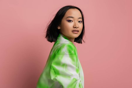 Photo for Brunette asian woman in stylish green and white jacket looking at camera isolated on pink - Royalty Free Image
