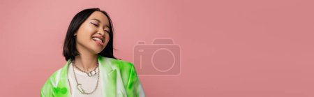 cheerful asian woman in green and white blazer sticking out tongue isolated on pink, banner