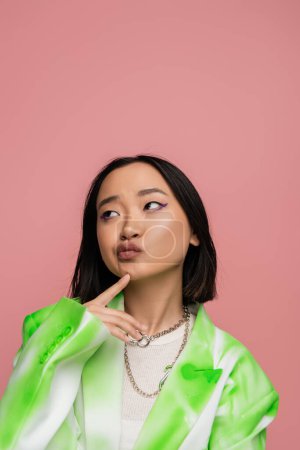 Photo for Thoughtful asian woman in trendy blazer and metal necklaces touching chin and looking away isolated on pink - Royalty Free Image