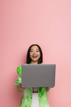 amazed asian woman in trendy clothes looking at camera near laptop isolated on pink
