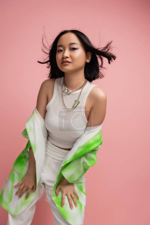 trendy asian woman in tank top and green and white blazer posing on wind isolated on pink