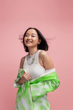 smiling asian woman in tank top and metal necklaces posing with green and white jacket isolated on pink