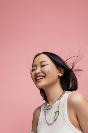 Foto de Low angle view of cheerful asian woman in white tank top and makeup with blue eyeliner isolated on pink - Imagen libre de derechos