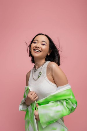 young and stylish asian woman in silver necklaces and white tank top looking at camera on wind isolated on pink