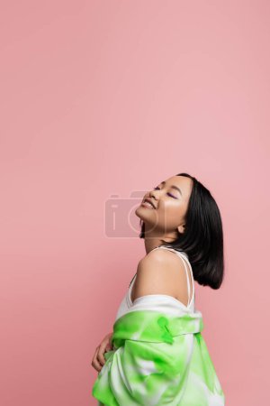 Photo for Pleased asian woman with closed eyes posing with green and white jacket isolated on pink - Royalty Free Image