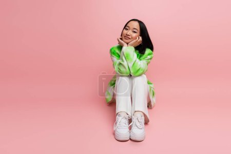 Photo for Full length of trendy asian woman in green and white outfit looking at camera while sitting on pink background - Royalty Free Image