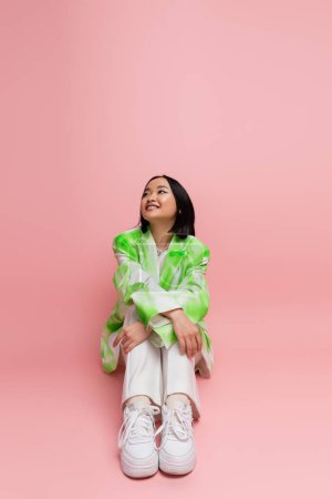 Photo for Full length of positive asian woman in green and white clothes sitting and looking away on pink background - Royalty Free Image