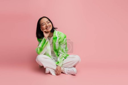 Téléchargez les photos : Pleased asian woman in fashionable outfit sitting with crossed legs on pink background - en image libre de droit