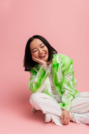 cheerful asian woman in green and white jacket sitting with crossed legs and sticking out tongue on pink background