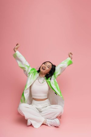 joyful asian woman in stylish outfit sitting with crossed legs and stretching raised hands on pink background