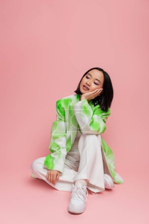 Photo for Pensive asian woman in white pants and trendy jacket sitting with hand near face on pink background - Royalty Free Image