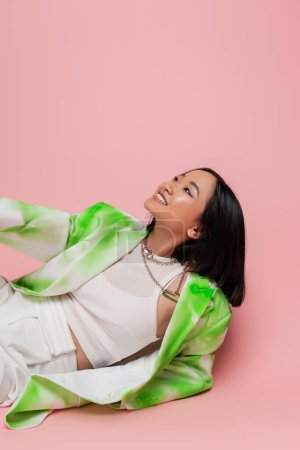 Photo for Happy asian woman in fashionable outfit and necklaces looking away while lying on pink background - Royalty Free Image