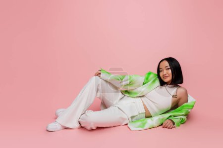Photo for Full length of smiling asian woman in white pants and fashionable jacket lying on pink background - Royalty Free Image