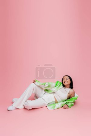 Photo for Full length of asian model in white pants and trendy jacket lying and looking at camera on pink background - Royalty Free Image