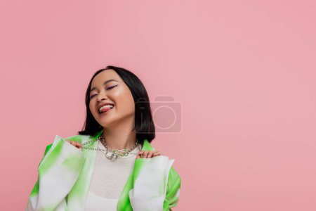 excited and flirty asian woman touching metal necklaces and sticking out tongue isolated on pink