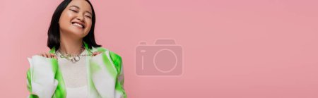 joyful asian woman in trendy outfit touching metal necklaces while posing with closed eyes on pink background, banner
