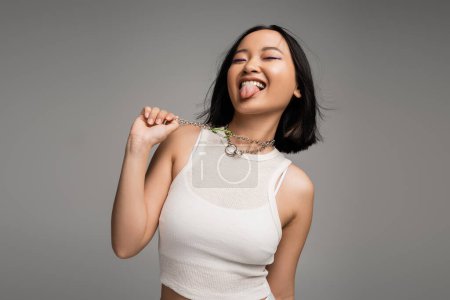 joyful asian woman in white tank top touching metal necklaces and sticking out tongue isolated on grey