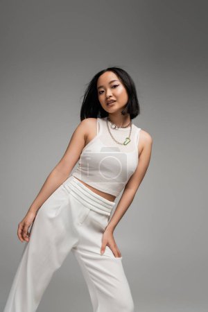 stylish asian woman in white pants and tank top with silver necklaces looking at camera isolated on grey