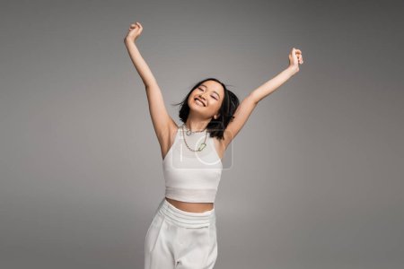 Photo for Pleased asian woman in white tank top and pants posing with closed eyes and raised hands isolated on grey - Royalty Free Image